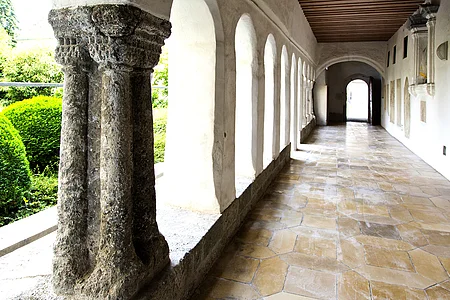 Steinbogen-Gang mit Säulen, der Kreuzgang des ehemaligen Klosters in Monheim, führt zu einer Tür, links grüne Büsche sichtbar. Entlang des Gangs alter Bodenbelag.