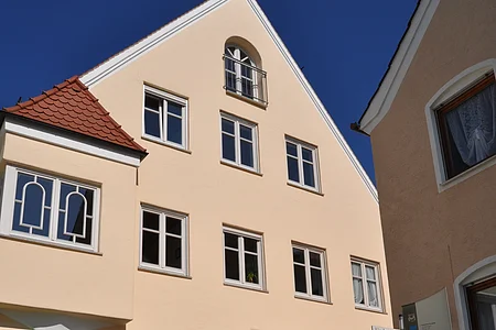 Ein dreistöckiges Gebäude mit Giebel, einem Türbogen und mehreren Fenstern auf einem gepflastertem Platz. Vor dem Gebäude steht ein Informationsschild und ein großer grauer Buchstabe "M" aus Stein. Der Buchstabe ist ein Teil des Monheimer Buchstabenweges.