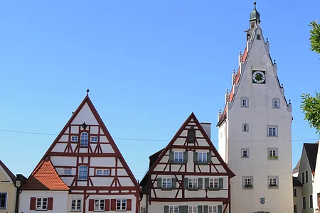 Historische Fachwerkhäuser, ein Torturm mit Uhr in einer Altstadt bei klarem Himmel. Links im Bild parkende Autos auf einem gepflasterten Platz. Rechts im Vordergrund ein Baum mit einer Info-Tafel und einem großen Buchstaben "O" aus Stein. Dahinter eine Informationsvetrine.