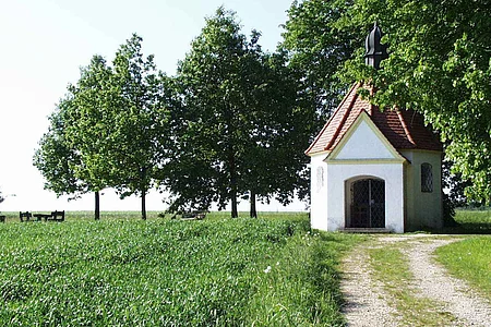 Kleine Kapelle mit rotem Dach auf einer Wiese mit einem Schotterweg. Umgeben von einem grünem Feld und Bäumen.
