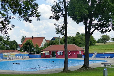 Ein Freibad mit Schwimmbecken, einer Rutsche und Sitzmöglichkeiten, umgeben von Bäumen und Wiesen. Im Hintergrund steht ein rotes Gebäude mit Fenstern, ein Kiosk.