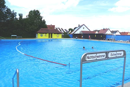 Ein Freibad mit einem großen Schwimmbecken, unterteilt in Nichtschwimmer- und Schwimmerbereich. Im Hintergrund sind Gebäude und Bäume zu sehen.