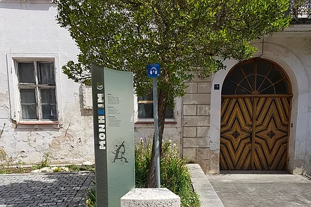 Steinskulptur des Buchstabens "E", dahinter ein Informationsschild und ein Baum mit einem dahinter liegendem Beet. Umgeben von einem gepflastertem Platz, mit einem historischen Gebäude im Hintergrund.