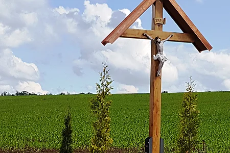 Holzkreuz mit überdachter Christusfigur mit bepflanztem Beet darunter, daneben eine Holzbank. Im Hintergrund ein Feld und blauer Himmel.