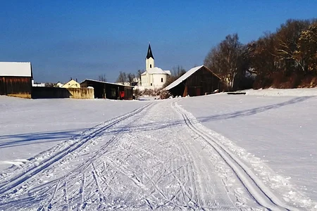 Verschneite Landschaft mit Skispuren, ein Ort zum Langlaufen in der Monheimer Alb. Im Hintergrund Gebäude, eine Kirche und blauer Himmel zu sehen.