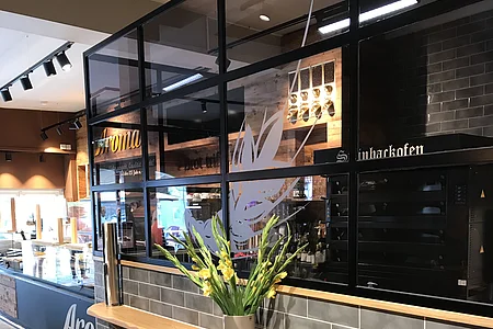 Innenraum eines modernen Cafés mit grauen Fliesen, einer Holzablage und einer Vase darauf mit gelben Blumen. Im Hintergrund ist eine Verkaufstheke und Küchengeräte.