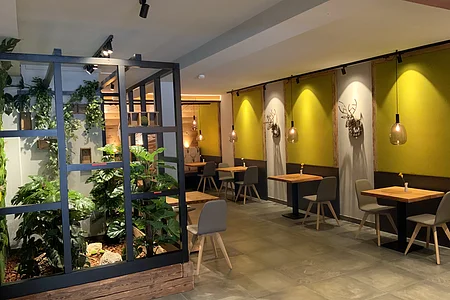 Innenraum eines modernen Cafés mit Holztischen, grauen Stühlen und Pflanzen hinter einer Glasfassade. An der Wand hängen beleuchtete Dekorationen in Form von Hirschgeweihen auf olivgrün gestrichenen Wänden.