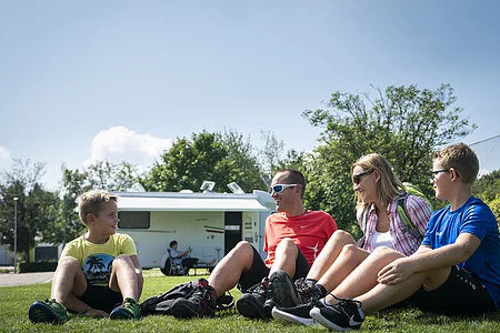 Eine Gruppe von vier Personen sitzt auf einer Wiese vor einem Wohnmobil und lacht.