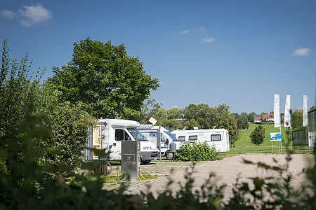 Wohnmobile parken auf einem Stellplatz, umgeben von Bäumen und Wiesen unter blauem Himmel.