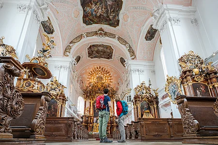 Zwei Personen mit Rucksäcken stehen in einer barocken Kirche mit verzierten Holzstühlen und bemalter Decke.