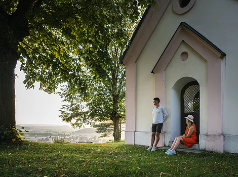 Wanderer auf der Monheimer Alb Mann lehnt an Hauswand, Frau sitzt in Türbogen, große Bäume, Dorf im Hintergrund bei Tageslicht.