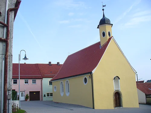 Gelbe Kapelle, die Peterskapelle in Monheim mit rotem Dach und einem Turm, umgeben von Wohnhäusern mit roten Dächern. Im Hintergrund blauer Himmel.