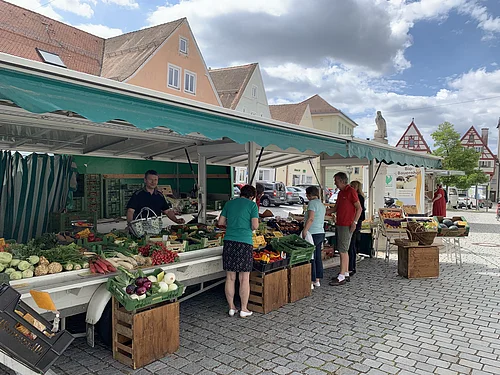 Marktstand des Wochenmarkts in Monheim mit Obst und Gemüse in der Auslage auf dem gepflastertem Marktplatz. Mehrere Personen kaufen dort ein. Fachwerkhäuser im Hintergrund und bewölkter Himmel.