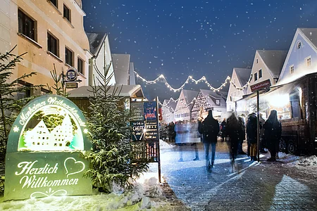 Monheimer Weihnachtsmarkt mit Verkaufsständen, Lichtern und Menschen. Ein beleuchtetes Schild links im Bild mit dem Text: "Monheimer Weihnachtsmarkt - Herzlich Willkommen" umgeben von mehreren Tannenbäumen. Schnee liegt auf dem gepflastertem Platz.