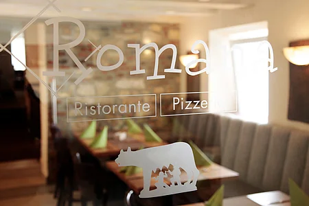 Fensterglas einer Pizzeria mit weißem Schriftzug „Romana Ristorante Pizzeria“ und Logo, dahinter unscharf zu sehen gedeckte Tische mit grünen Servietten und einer Steinwand.