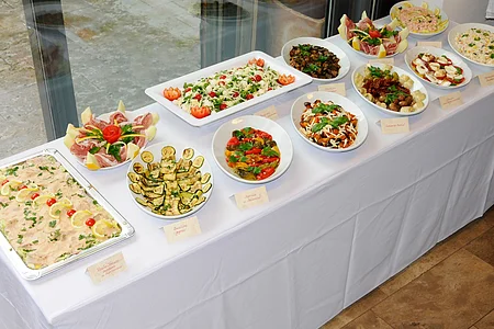 Buffet mit vielen verschiedenen Salaten und Antipasti mit kleinen Schildern daneben auf einem weiß gedecktem Tisch vor einem Glasfenster.