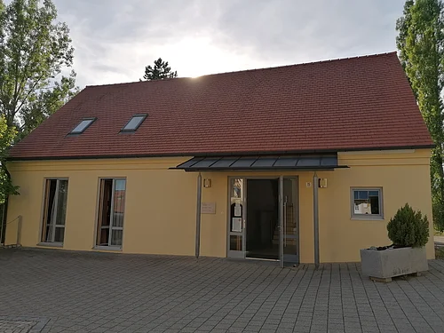 Das Haus Martin Luther in Monheim, ein evangelisches Gemeindehaus: Ein gelbes Gebäude mit rotem Satteldach, zwei Fenstern und einer offenen Eingangstür, umgeben von Bäumen. Vor dem Haus steht ein bepflanztes Beet.