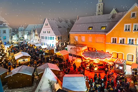 Der Monheimer Weihnachtsmarkt mit zahlreichen Besuchern, festlich beleuchteten Ständen und Kinderkarussell bei Schnee in historischen Altstadt von Monheim.