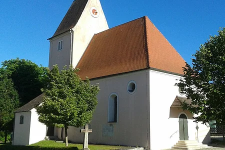 Kirche mit rotem Satteldach und einer Turmuhr umgeben von Bäumen bei klarem Himmel, an einem gepflastertem Weg.