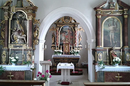 Innenraum einer Kirche mit Altar, zwei Seitenaltären mit Heiligenfiguren und Blumenarrangements an einer weißer Wand und mit einem Durchgangs-Bogen gestaltet.