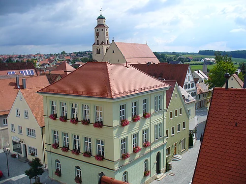 Blick von oben auf eine Innenstadt mit Hauptaugenmerk auf ein historisches grünes Gebäude im Vordergrund, mit roten Dachziegeln und Blumen an den Fenstern.  Im Hintergrund weitere Häuser der Kleinstadt und blauer Himmel. Rechts unten im Eck das rote Dach eines anderen Gebäudes im Vordergrund zu sehen.