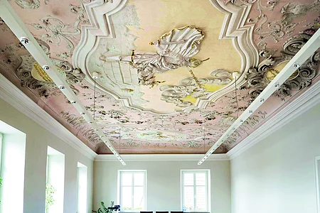 Decke mit barocker Stuck- und Freskoverzierung in einem hellen Raum mit Fenstern und moderner Beleuchtung.