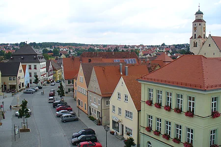 Blick auf eine Straße in der Monheimer Innenstadt mit geparkten Autos an der Straße entlang und Fachwerkhäusern mit roten Dächern bei bewölktem Himmel. An der Straße einzelne Bäume gepflanzt, ein Brunnen auf der linken Seite ist zu sehen und auf der rechten Seite eine Kirche.