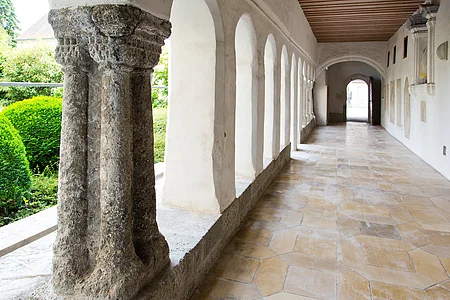 Blick in einen langen Kreuzgang mit Säulen und Rundbögen links im Bild, dahinter grüne Büsche. Rechts im Bild weiße Wand mit einer Ausfräsung in der Wand und Steinplatten mit Text angebracht. Bodenbelag aus hellen alten Steinen.