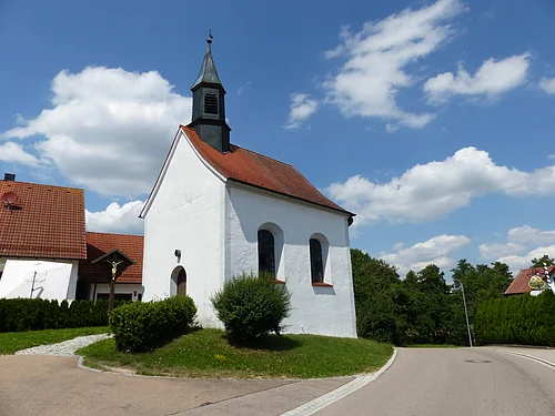 Kleine weiße Filialkirche mit rotem Dach und kleinem Turm oben drauf an einer Straße bei blauem Himmel mit Wolken. Umgeben von grüner Wiese und Hecken, daneben ein Jesuskreuz aus Holz. Im Hintergrund weiteres weißes Haus zu sehen.
