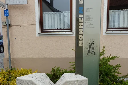 Steinerner grauer Buchstabe M vor Infotafel aus Glas mit Erklärtext und einem Stadtplan. Auf der Infotafel vertikal der Text „Monheim“. Im Hintergrund ein Haus in beige mit einer Hecke und Straßenschildern.