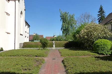 Ehemaliger Klostergarten in Monheim mit gepflastertem Weg in der Mitte, niedrigen Hecken, Büschen und Bäumen. Am Ende des Weges eine Statue vor einer Hecke unter blauem Himmel. Links im Bild ein weißes Gebäude.
