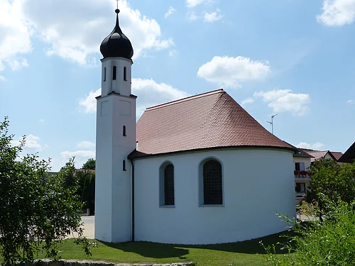 Weißes Kirchgebäude mit abgerundetem Grundriss und kleinem Turm mit schwarzer Haube bei blauem Himmel, umgeben von grünen Bäumen und wiese. Im Hintergrund weitere Häuser.