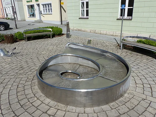 Juraschneck-Brunnen Monheim Juraschneck-Brunnen: ein Brunnen aus Metall in Form eines Fossils einer Schnecke, mit eingravierten Texten auf der Oberfläche. Außenrum ein gepflastertem Platz mit zwei Sitzbänken. Im Hintergrund farbige Häuser und eine kleine grüne Hecke an den Bänken entlang.