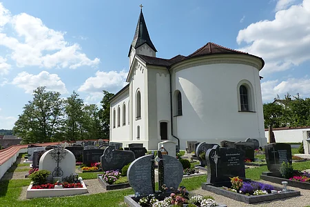 Weiße Pfarrkirche mit abgerundetem Gebäude, dunklem Dach und einem kleinen Turm mit schwarzem Dach. Anliegend ein Friedhof  mit Grabbepflanzung und Grabsteinen bei sonnigem Himmel und wenigen Wolken.