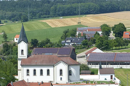 Pfarrkirche mit abgerundetem Gebäude und einem Turm, Felder und Wald im Hintergrund an einem sonnigen Tag. Umliegende Häuser mit Solarpaneelen.