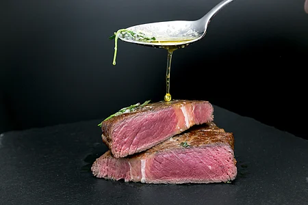 Ein Löffel gießt Öl über zwei aufeinander gestapelte, halbierte Rindersteaks auf einem schwarzen Hintergrund.