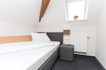 Einzelzimmer in einem Hotel mit einem Einzelbett links an der weißen Wand, einem grauem Hocker rechts daneben, auf dem eine  Fernbedienung liegt und einer grauen Leuchte oberhalb an der Wand. Dachschräge mit Holzbalken, rechts davon und kleines Fenster mit Pflanze auf dem Fensterbrett. Unter dem Fester eine kleine weiße Heizung an der Wand angebracht.