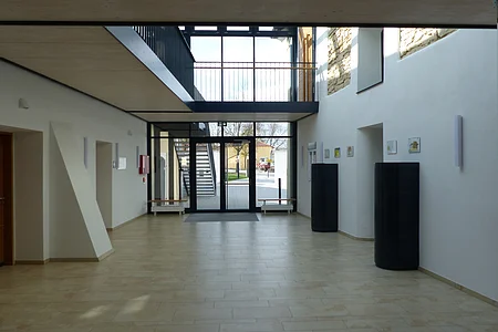 Heller Eingangsbereich eines großen Raums mit Ausgang nach hinten ins Freie, Ausgangstür bestehend aus Glas. Zwei schwarze Säulen an der Wand rechts im Bild. Zwei Heizkörpern mit einer Sitzauflage darauf und einem offenem Obergeschoss mit schwarzem Metallgeländer darüber. Im Freien links im Bild eine schwarze Treppe führend in das Obergeschoss zu sehen.