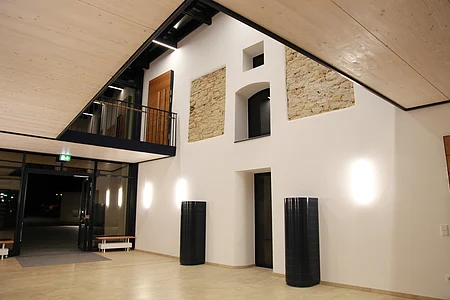 Heller Innenraum mit heller verkleideter Holzdecke, weißen Wänden und hellem Bodenbelag. Zwei schwarze Säulen rechts an der Wand und offener Glastür bei Nacht. Oben im offenem Obergeschoss zwei Teilbereiche einer freigelegten Steinmauer in der Wand, eine braue Holztür und schwarzes Metallgeländer.