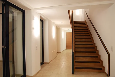 Flur mit beleuchteten weißen Wänden durch Wandleuchten links im Bild und einer Glastür. Eine Holztreppe, die nach oben führt rechts im Bild. Hinten im Gang eine helle Holztür, heller Bodenbelag aus Fließen.