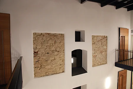 Innenraum mit weißer Wand, darin zwei Ausschnitte einer freigelegten Steinmauern und mehreren hellen Türen aus Holz mit schwarzen Metallgeländern. Oben an der Decke dunkle Holzbalken zu sehen.