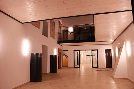 Beleuchteter heller Innenraum mit heller Holzdecke, weißen Wänden, zwei schwarzen Säulen links an der Wand, einer mit Holz verkleideten Treppe und mehreren Türen und Fenstern. Heller Bodenbelag aus Fließen.