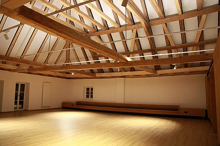 Beleuchteter leerer Raum mit offener Holzbalkendecke, hellem Holzboden und weißen Wänden, Fenster, Tür und eine Heizung an der Wand auf der linken Seite.