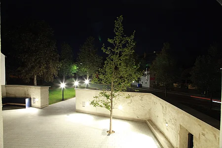 Beleuchteter Innenhof bei Nacht, eingezäunt mit einer hohe weißen Mauer, darin ein Baum rechts im Bild und eine an der Wand befestigte Sitzbank links im Bild. Außerhalb drei leuchtende Laternen am Gehweg entlang, Bäume auf grüner Wiese und im Hintergrund eine Straße mit einem grauen Haus.