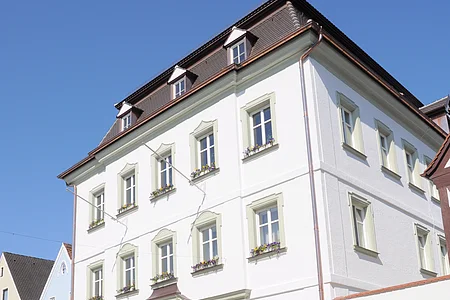 Das Rathaus der Stadt Monheim, am Marktplatz. Ein weißes, dreistöckiges Gebäude mit Satteldach und mehreren Fenstern, im Vordergrund gepflasterter Marktplatz.