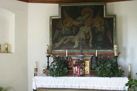 Innenansicht einer Kapelle: Altar mit weißem Tuch bedeckt, umgeben von verschiedenen Kerzen, Blumen und religiösem Gemälde oberhalb an der Wand, in einem Raum mit Holzbalkendecke.