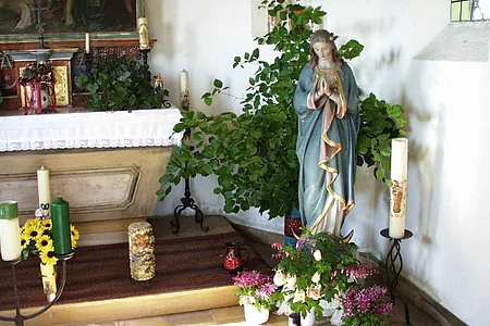 Innenansicht einer kleinen Kapelle mit der Statue einer betenden Frau mit blauem Mantel in einer Ecke umgeben von Kerzen, Pflanzen und Blumen in einem weißen Raum. Links im Bild ein geschmückter Altar.