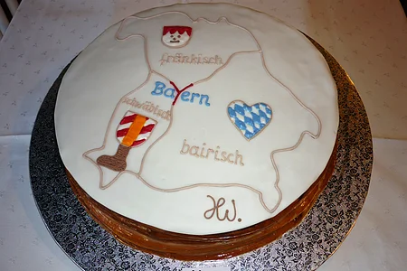 Runde Torte auf einer silbernen Unterlage seht auf einem Tisch mit weißem Tischtuch. Auf der Torte weiße Glasur und eine Karte von Bayern mit Dialektbezeichnungen und den dazugehörigen Symbolen darauf.