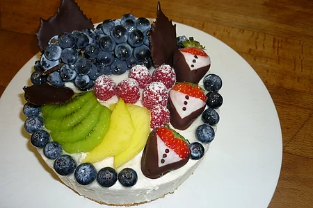 Obsttorte mit weißer Creme, obendrauf Trauben, gezuckerten Himbeeren, Kiwi, Ananas, Blaubeeren und Erdbeeren in Schokoladenmantel auf einem weißem Teller serviert.