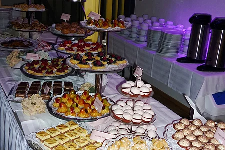 Buffet mit verschiedenen süßen Kuchen, Gebäck und Obsttörtchen auf mehreren Etageren und Tellern auf einem Tisch aufgedeckt. Im Hintergrund sind aufgestapelte beleuchtete Teller und Tassen mit zwei Kaffeekochern.